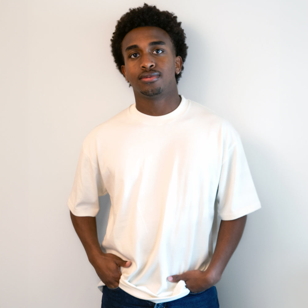 A man wearing a plain white short-sleeve crewneck t-shirt and dark blue jeans, standing against a light gray wall.