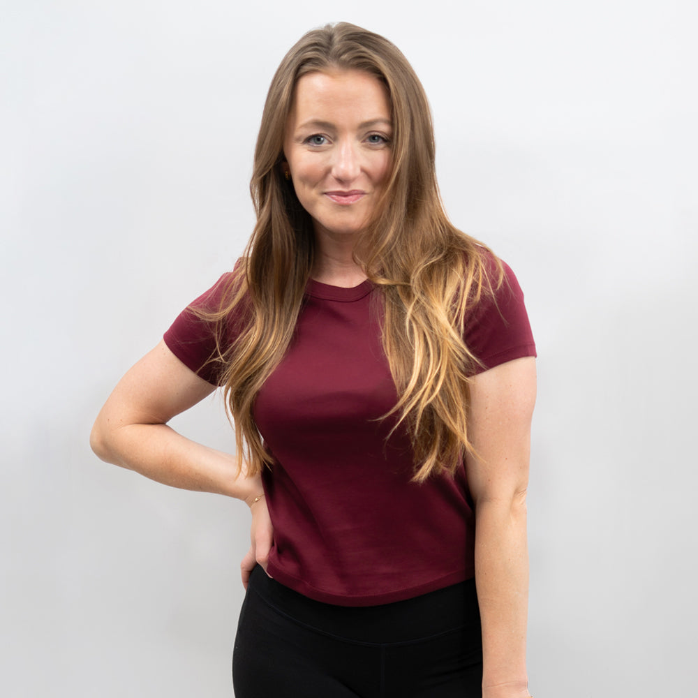 A woman with long light brown hair wearing a short-sleeve crewneck t-shirt in burgundy and black pants, standing against a light gray studio background.