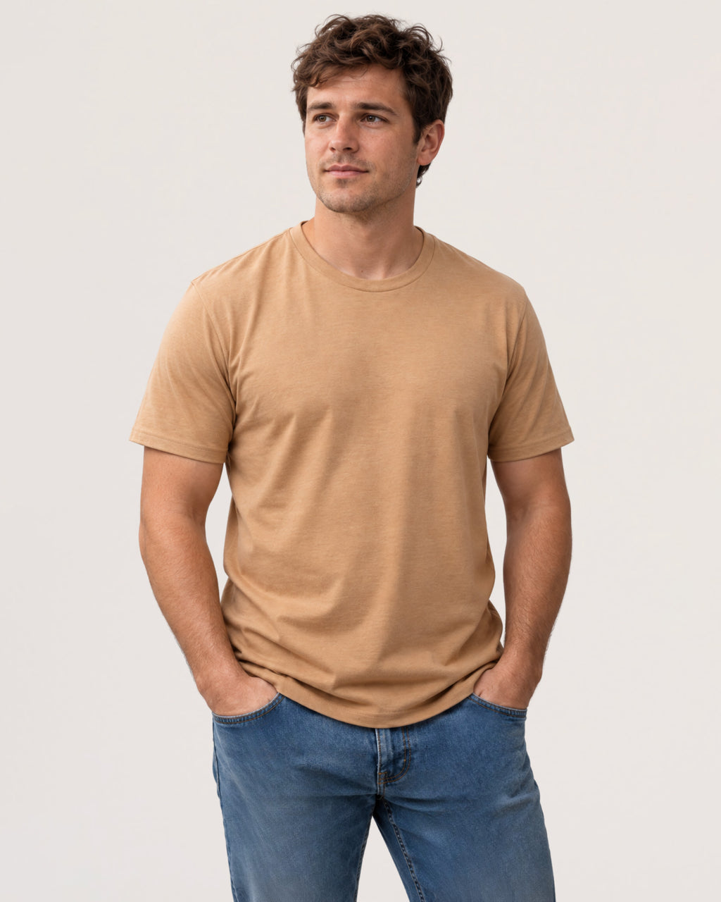 A man wearing a light tan crew-neck t-shirt and blue jeans, standing against a plain light gray studio background.