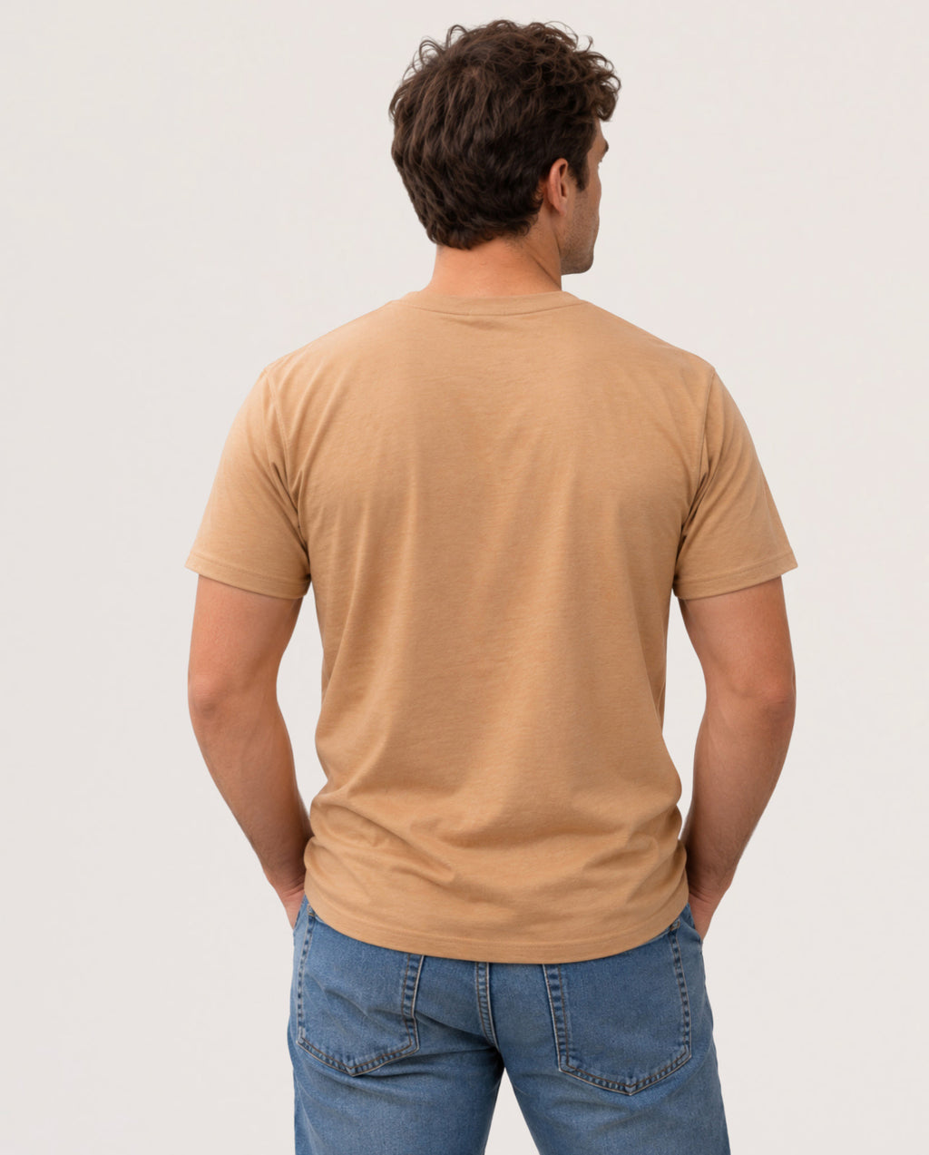 A man viewed from behind wearing a plain tan crew-neck t-shirt and blue jeans, standing against a light gray studio background.