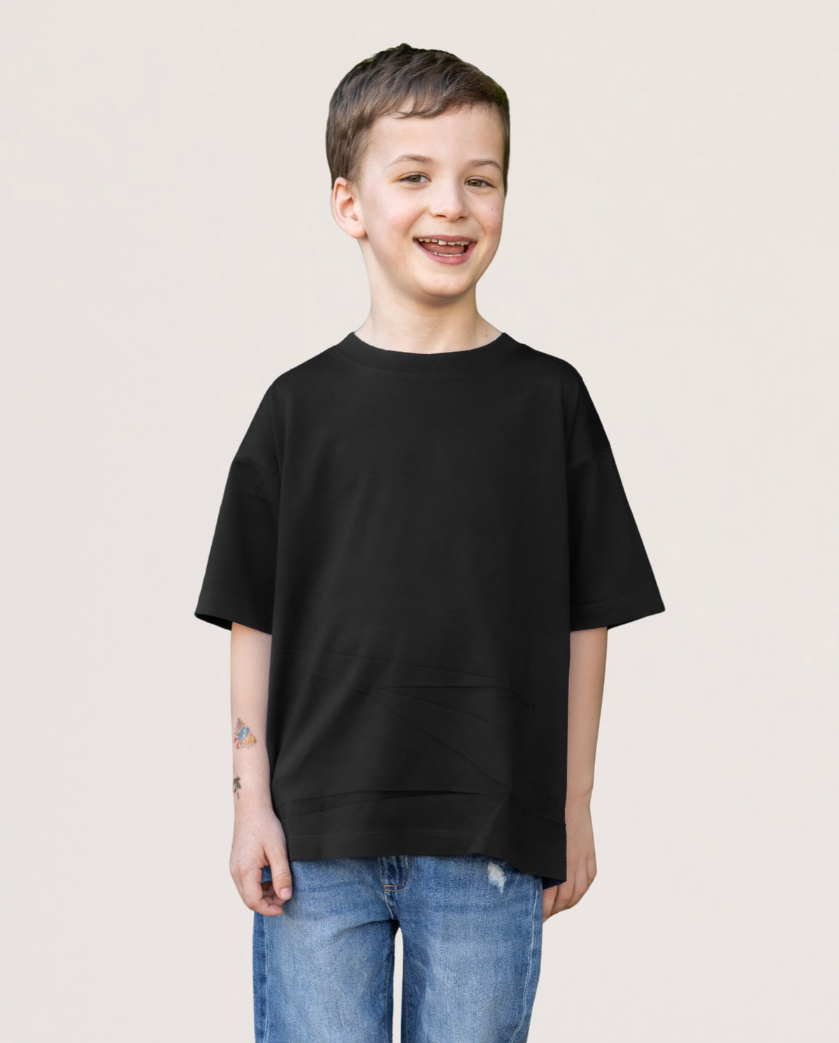 A young boy smiling and wearing an oversized black short-sleeve t-shirt with side pockets, paired with blue jeans, against a plain light beige background.