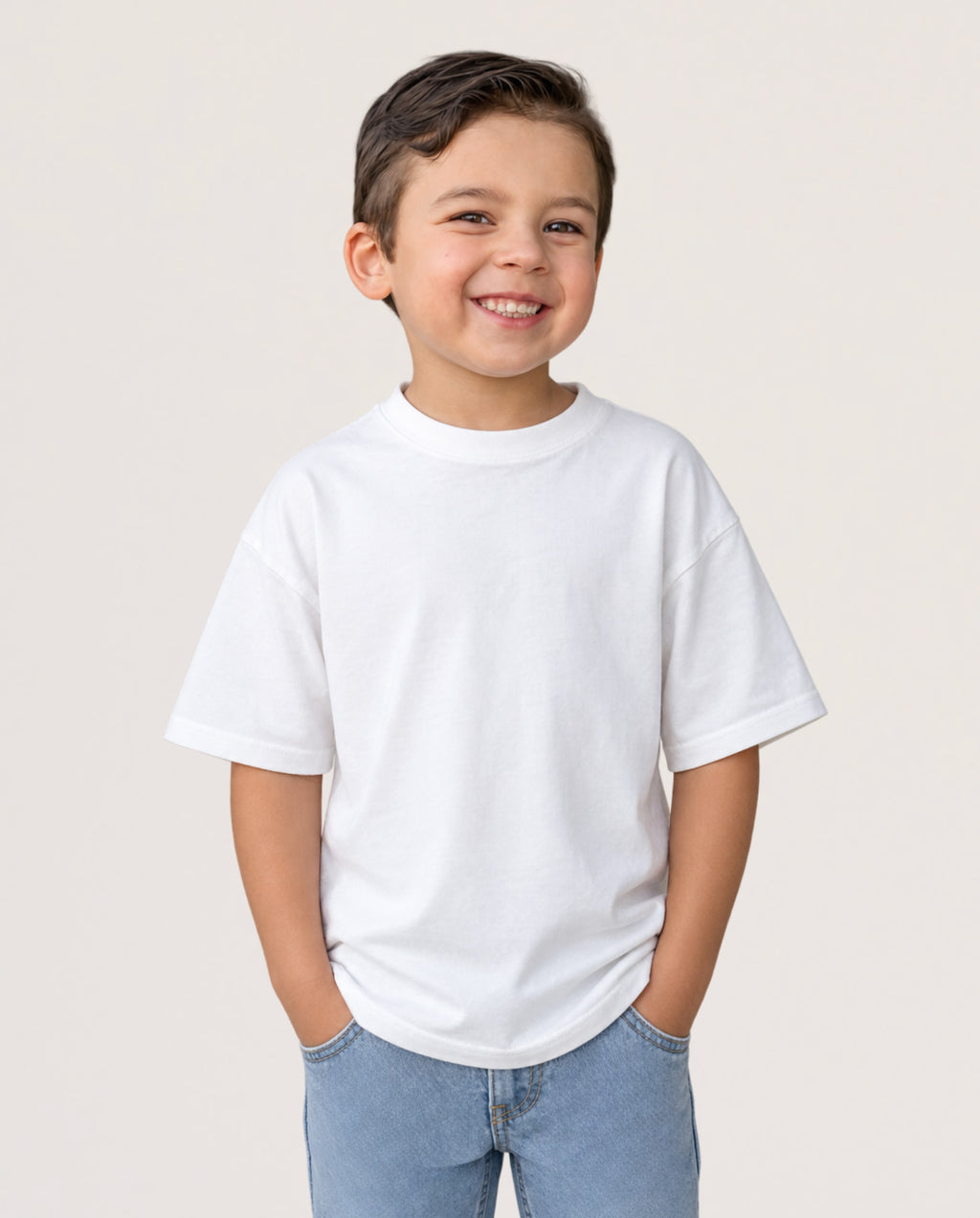 A young boy smiling and wearing a plain white short-sleeve crewneck t-shirt with light blue jeans, standing against a solid light beige background.