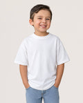 A young boy smiling and wearing a plain white short-sleeve crewneck t-shirt with light blue jeans, standing against a solid light beige background.