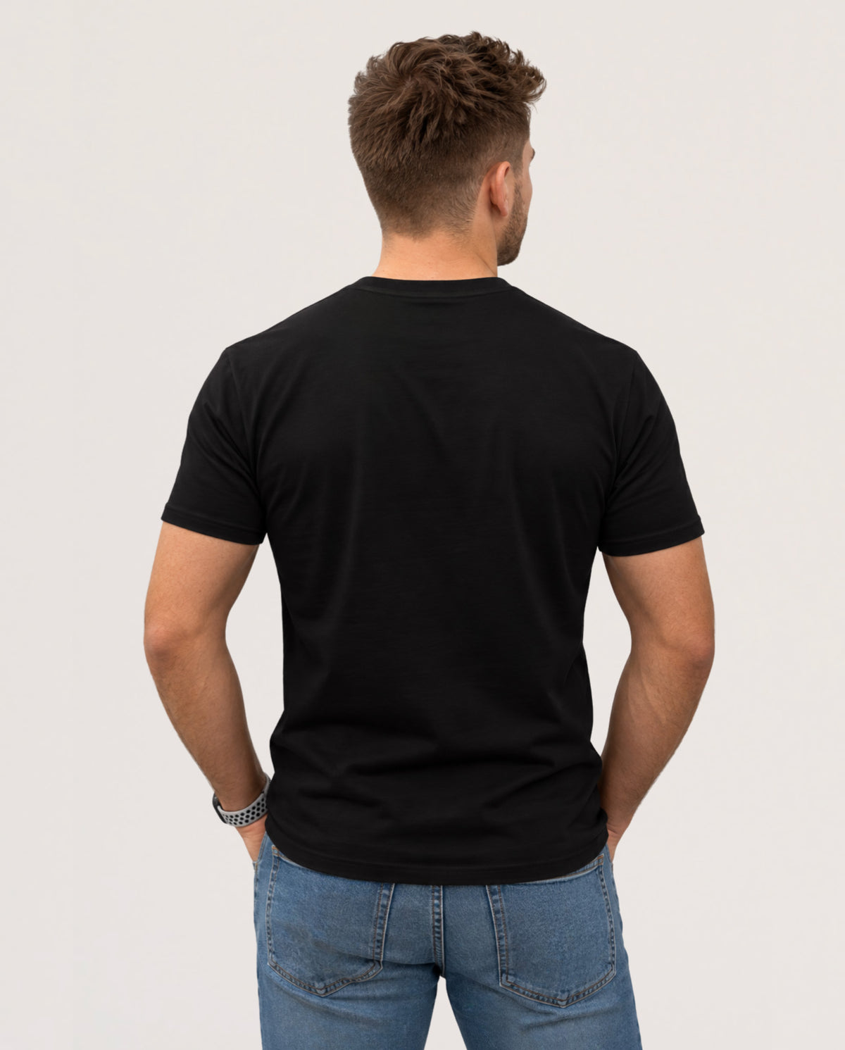 A man viewed from behind wearing a plain black short-sleeve t-shirt and blue jeans, standing against a light gray studio background.