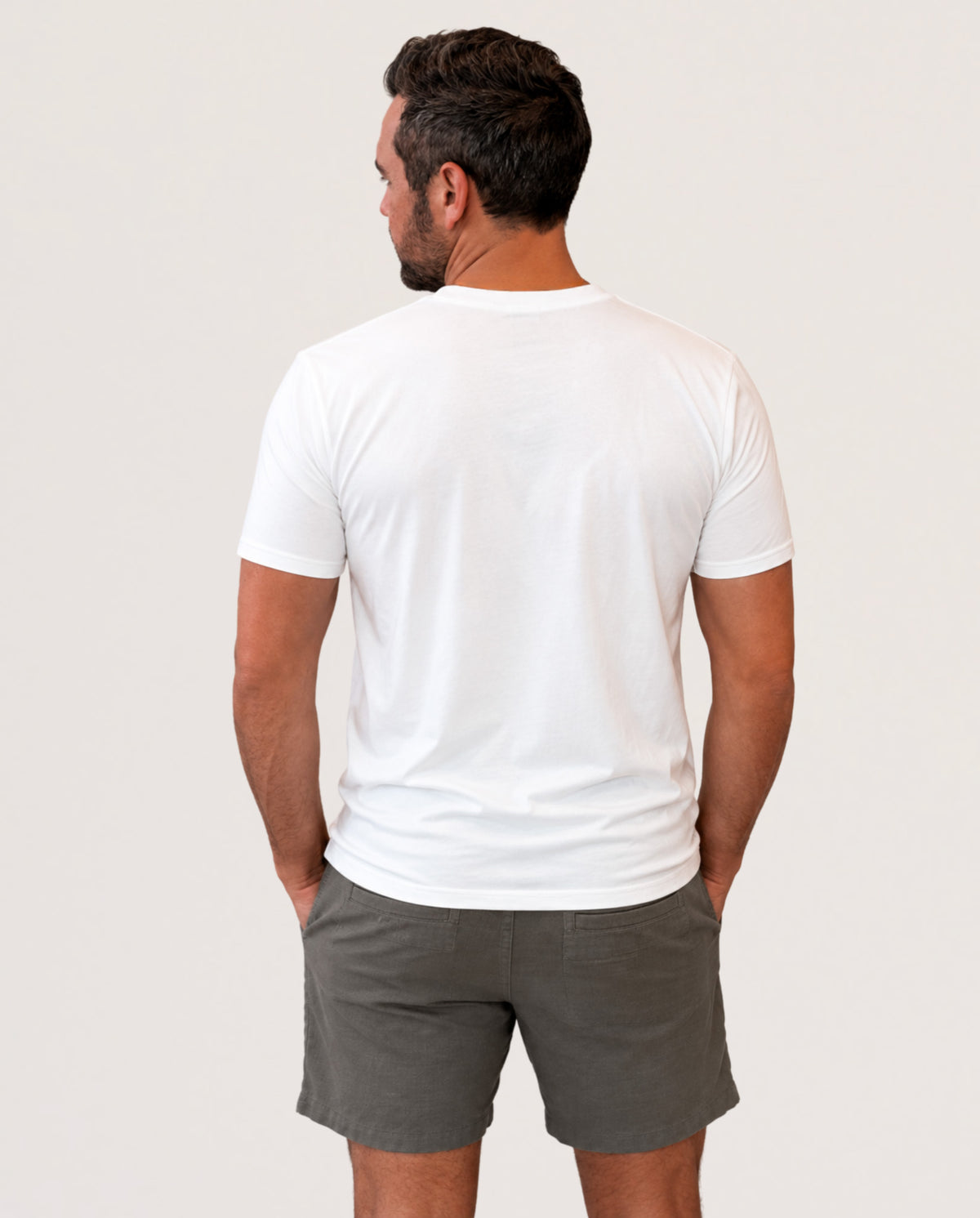 A man viewed from behind wearing a plain white short-sleeve crewneck t-shirt and dark gray shorts, standing against a light off-white studio background.
