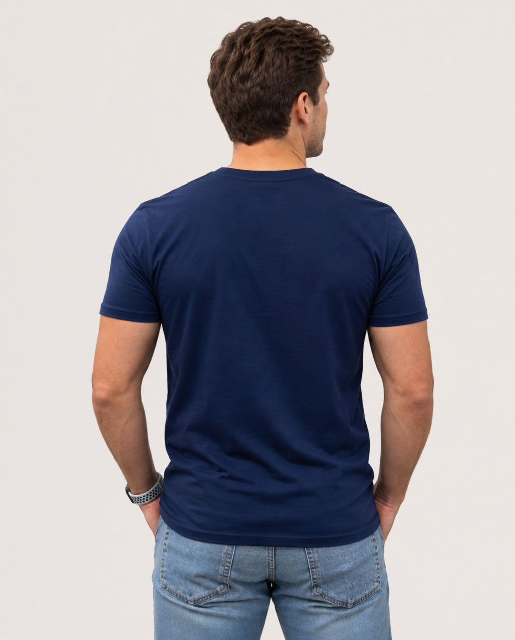 A man viewed from behind wearing a navy blue short-sleeve crewneck t-shirt and light blue jeans, standing against a plain light gray background.