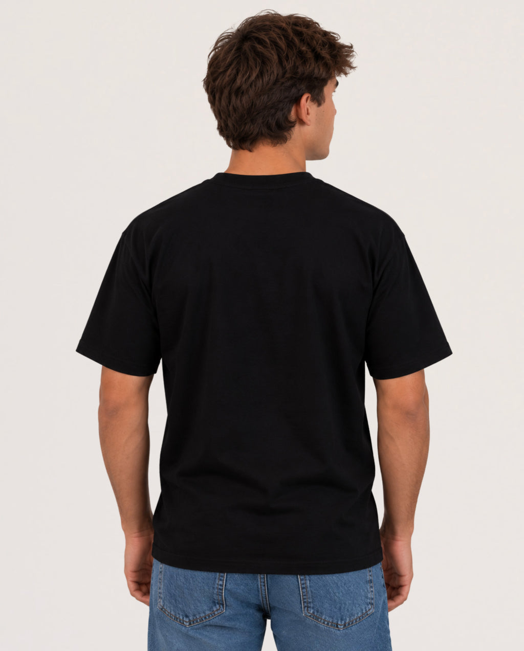 A man viewed from the back wearing a plain black short-sleeve crewneck t-shirt and blue jeans, standing against a light gray studio background.
