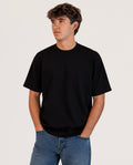 A young man wearing a plain black short-sleeve crewneck t-shirt and blue jeans, standing against a light gray studio background.