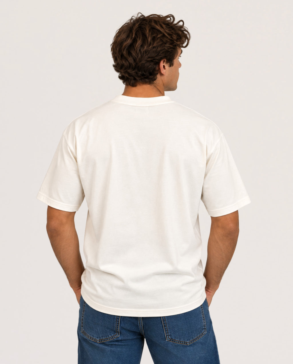 A man viewed from behind wearing a plain off-white short-sleeve t-shirt and blue jeans, standing against a light gray studio background.