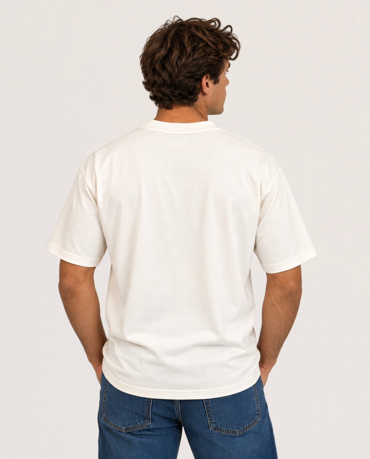 A man viewed from behind wearing a plain off-white short-sleeve t-shirt and blue jeans, standing against a light gray studio background.