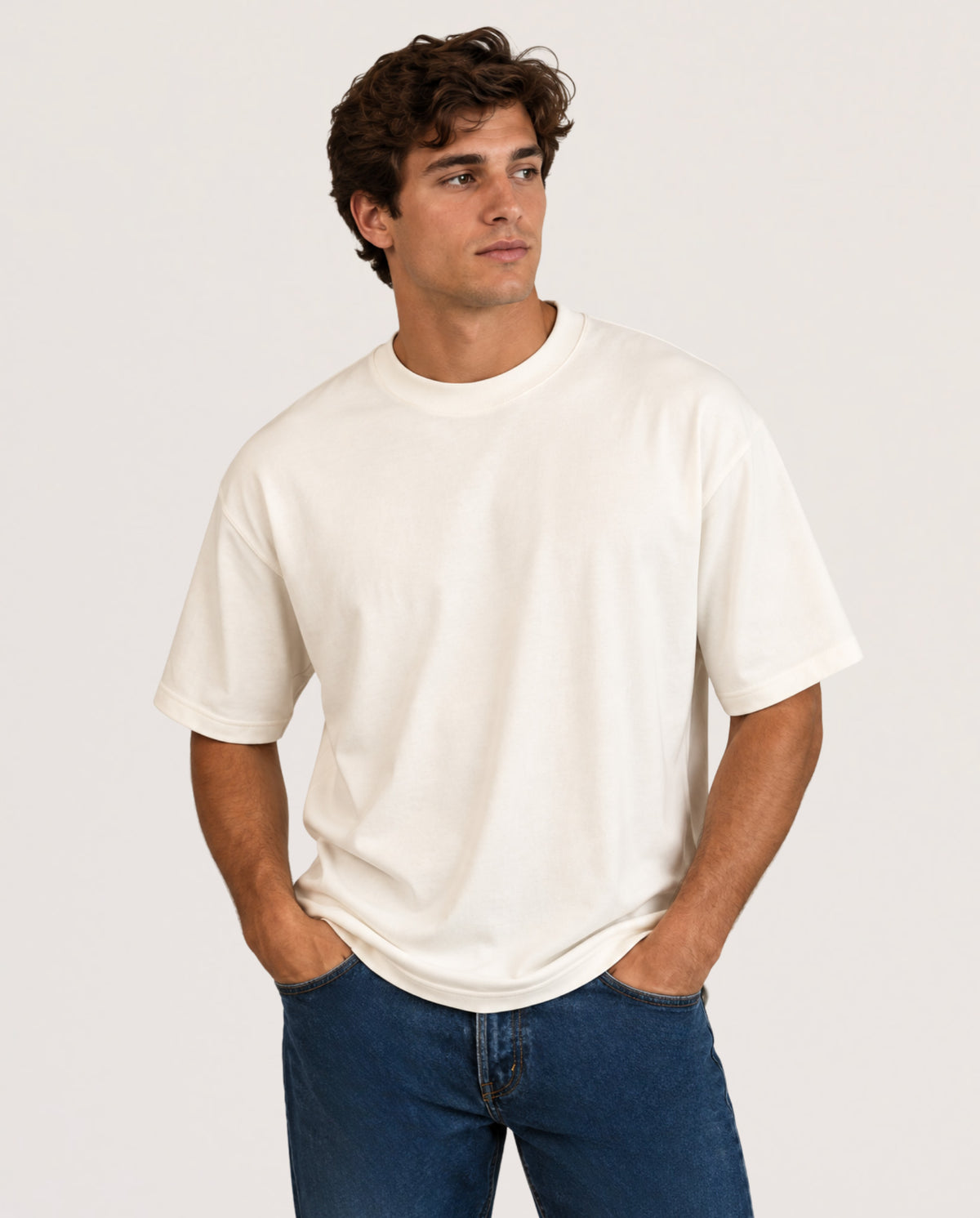 A man wearing a plain off-white short-sleeve crewneck t-shirt and blue jeans, standing against a light gray studio background.