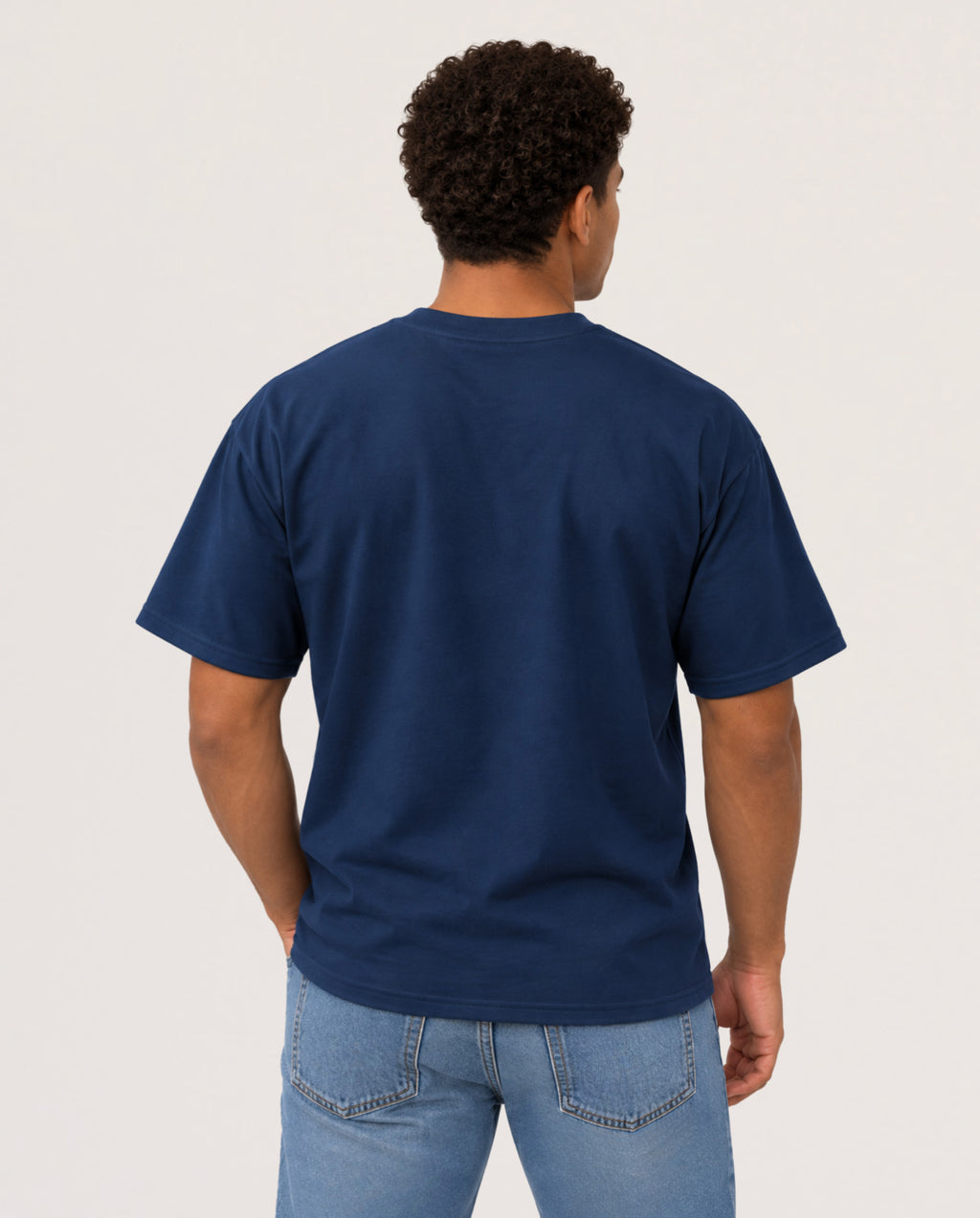 A man viewed from behind wearing a navy blue short-sleeve t-shirt and light blue jeans, standing against a plain light gray studio background.