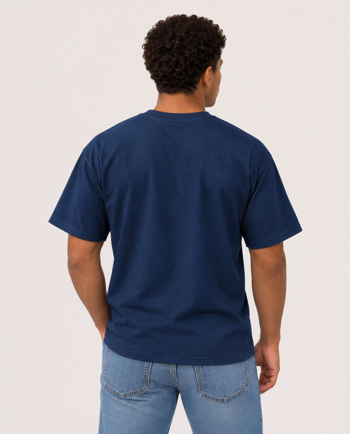 A man viewed from behind wearing a navy blue short-sleeve t-shirt and light blue jeans, standing against a plain light gray studio background.