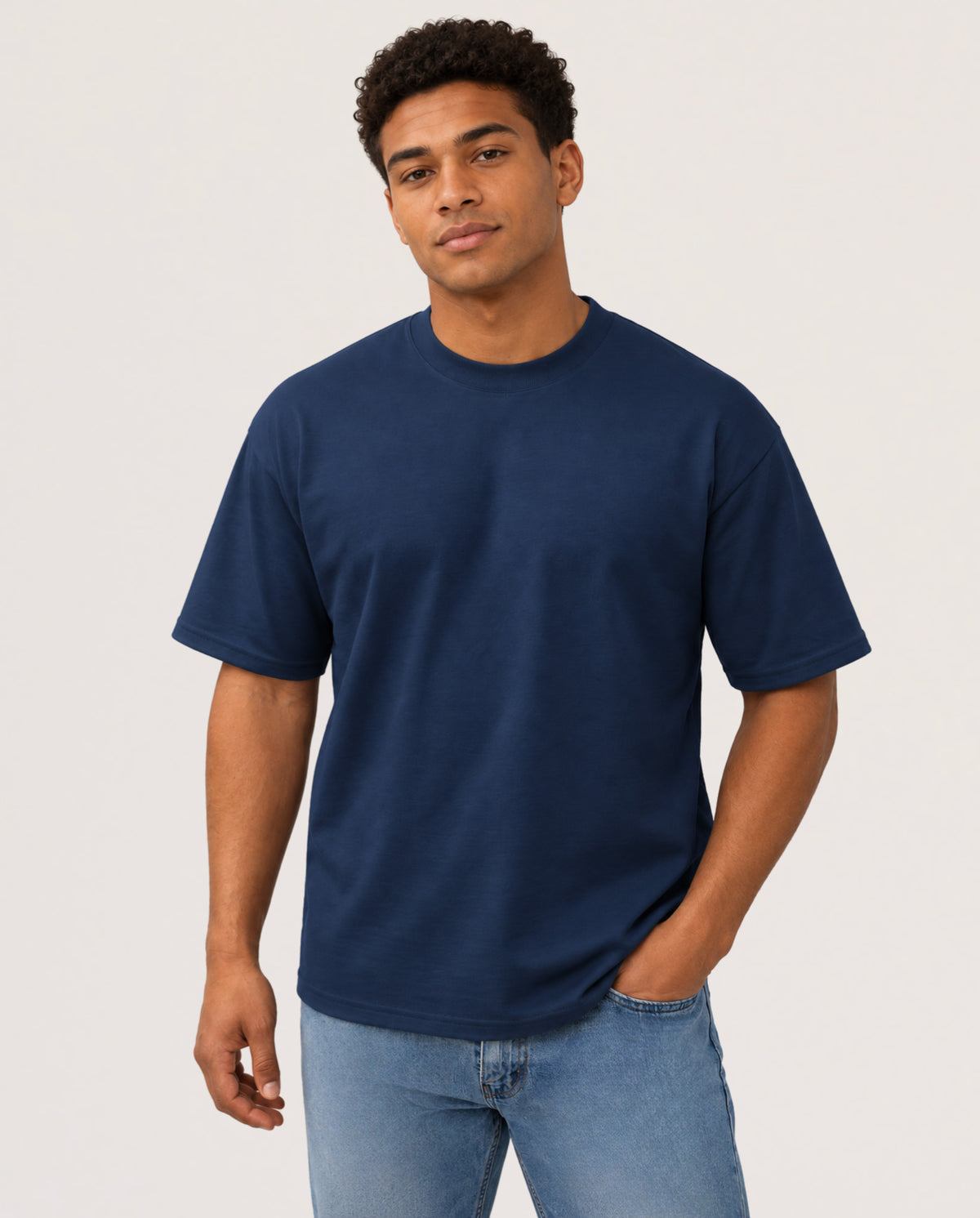 A man wearing a navy blue crewneck t-shirt and light blue jeans, standing against a plain light gray studio background.