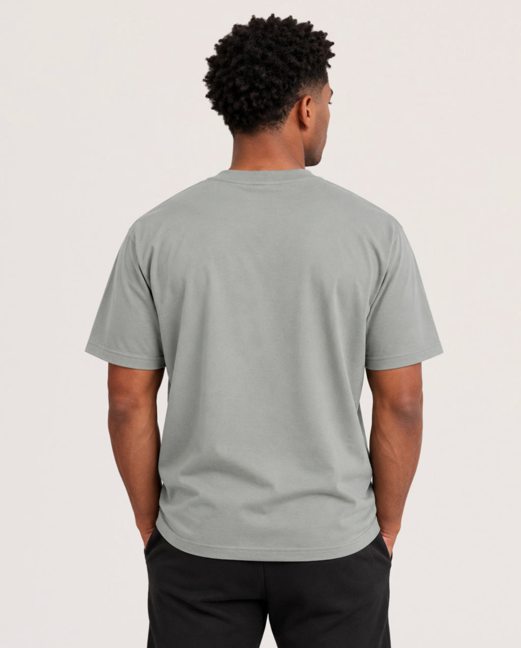 A man viewed from behind wearing a light sage green crewneck t-shirt and black pants, standing against a plain off-white studio background.
