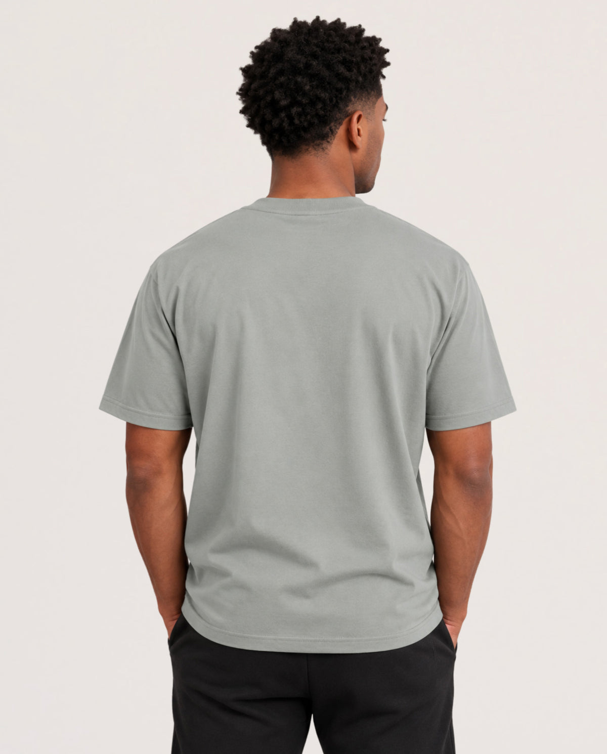 A man viewed from behind wearing a light sage green crewneck t-shirt and black pants, standing against a plain off-white studio background.