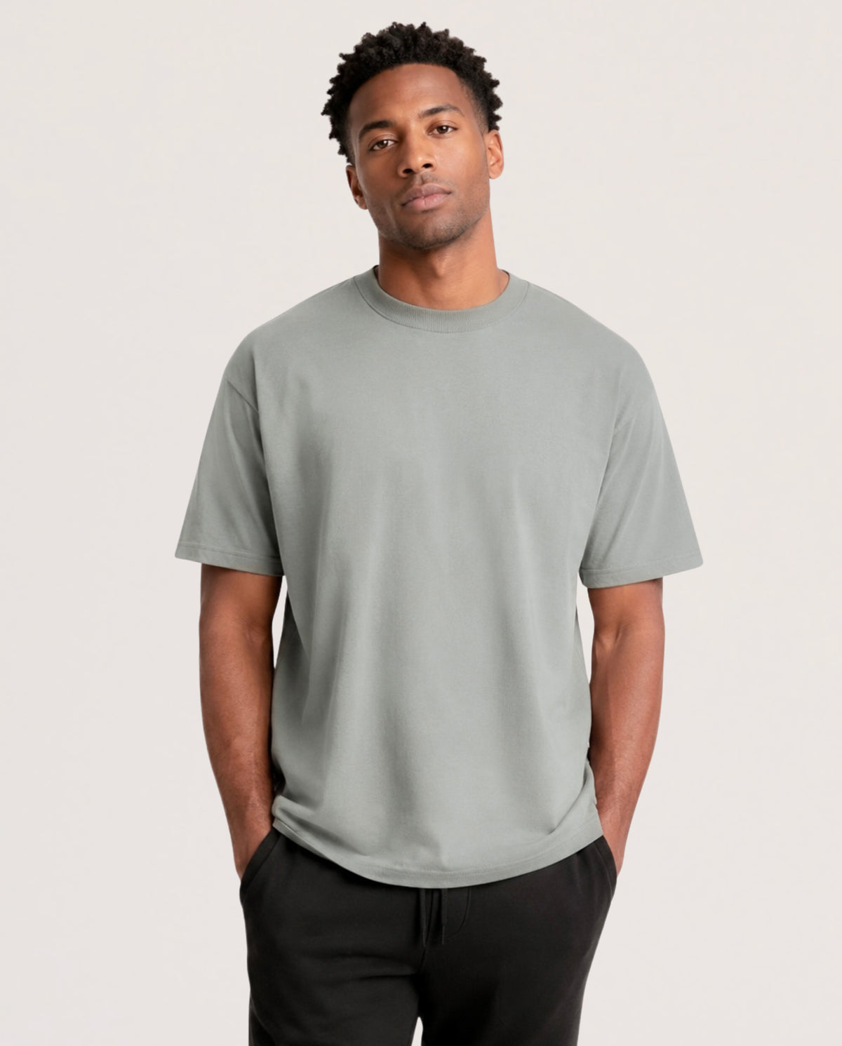 A man wearing a light sage green crewneck t-shirt and black sweatpants, standing against a plain light gray studio background.