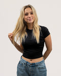 A blonde woman wearing a fitted black short-sleeve crop top and blue jeans, standing against a plain light gray studio background.