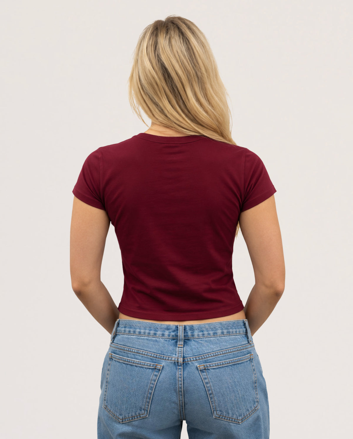 A woman viewed from behind wearing a fitted short-sleeve crewneck t-shirt in burgundy and light wash blue jeans, set against a plain off-white studio background.