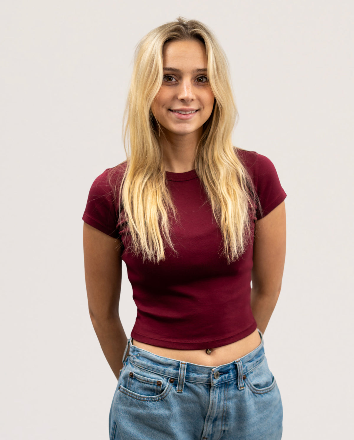 A blonde woman wearing a fitted short-sleeve crop top in deep burgundy and light wash blue jeans, standing against a plain light gray studio background.