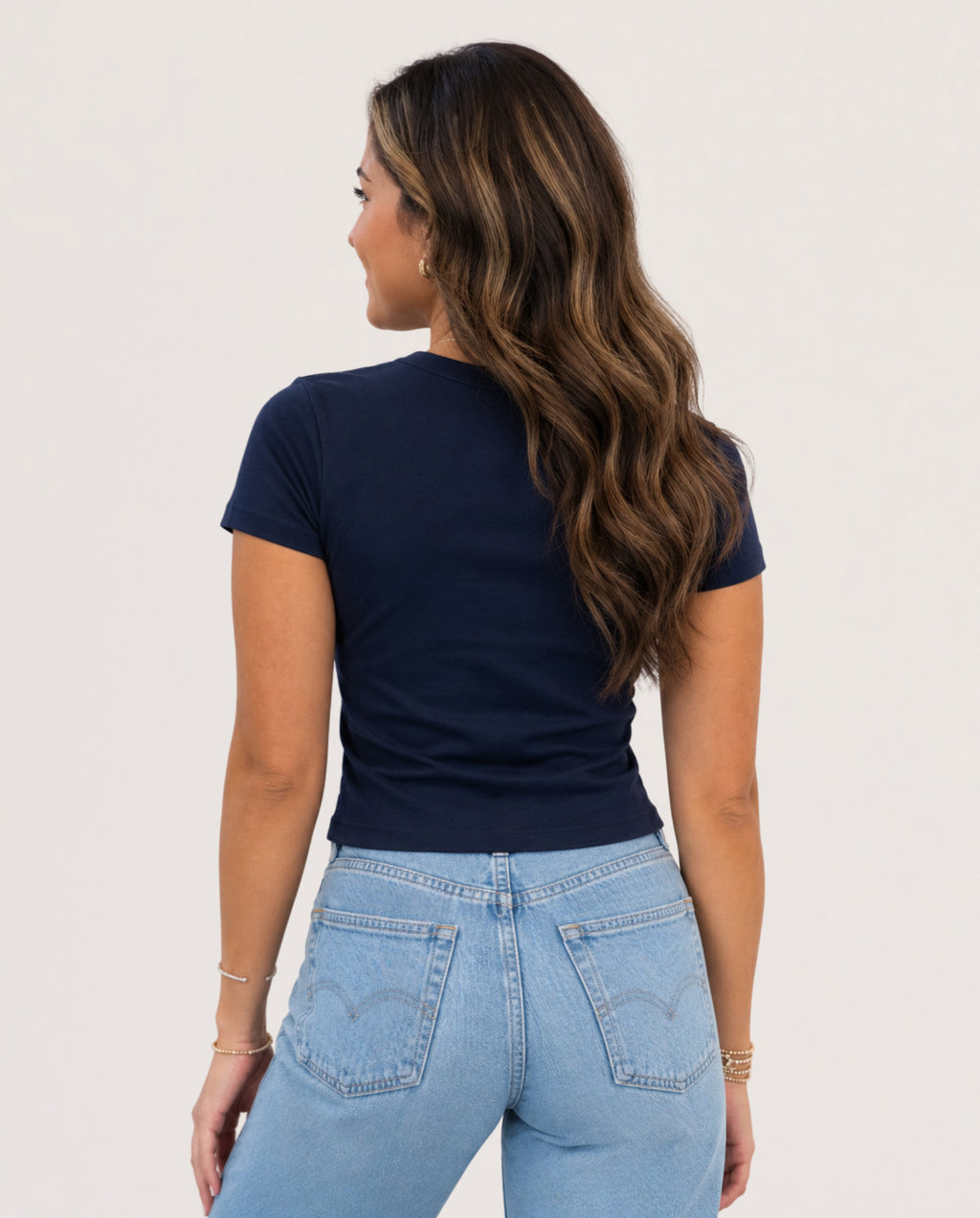 A woman shown from the back wearing a navy blue short-sleeve crewneck t-shirt and light wash blue jeans against a plain off-white background.