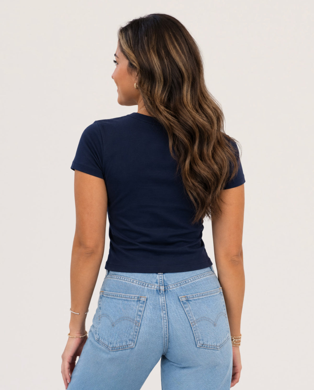 A woman shown from the back wearing a navy blue short-sleeve crewneck t-shirt and light wash blue jeans against a plain off-white background.