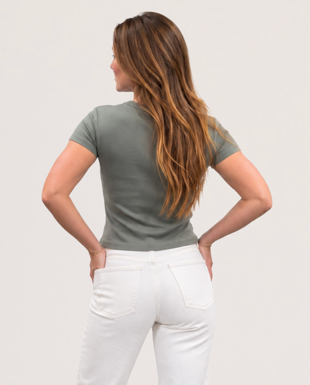 A woman with long brown hair is shown from behind wearing a fitted short-sleeve t-shirt in sage green and white pants, posing with her hands on her hips against a plain light gray background.