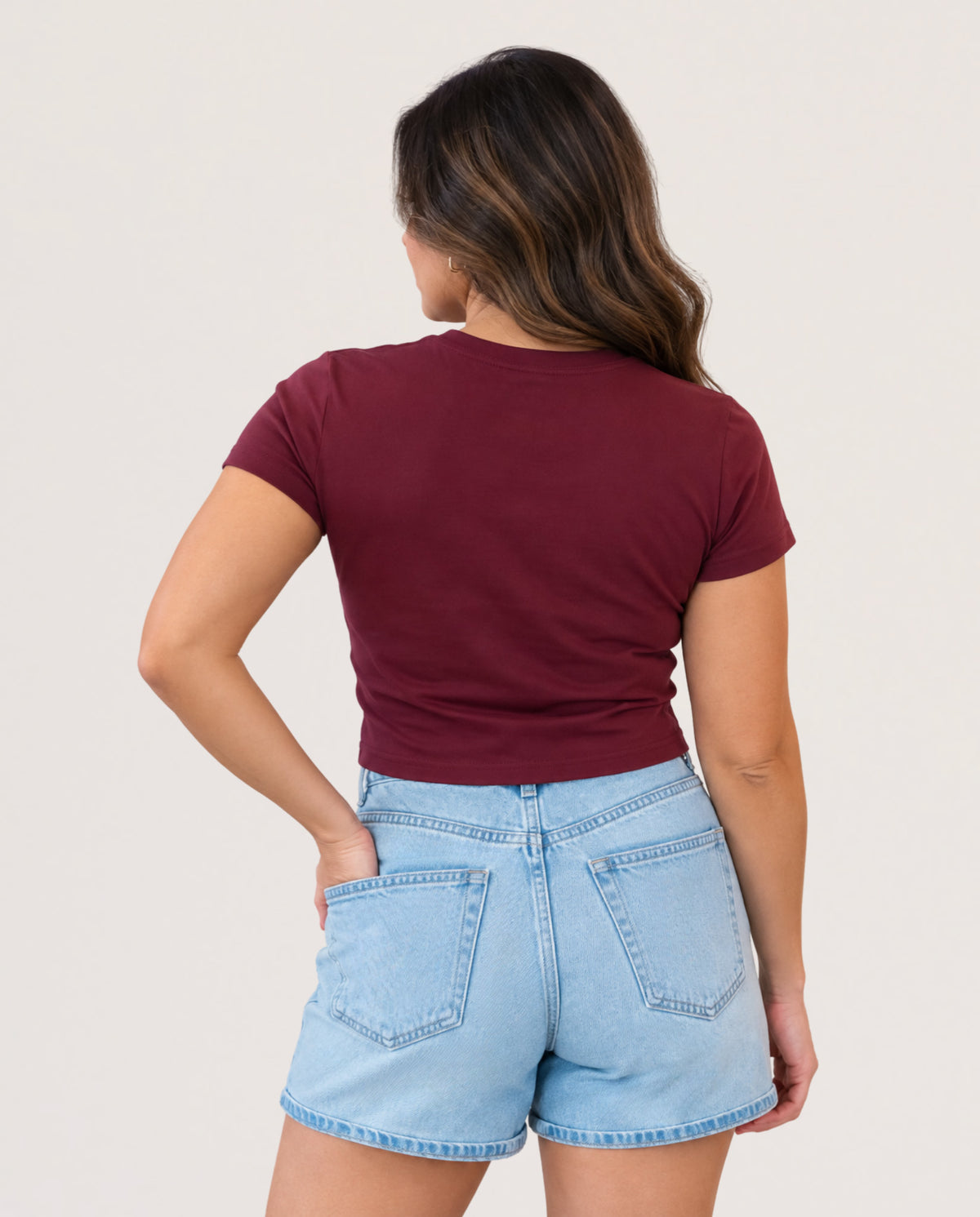 A woman shown from the back wearing a fitted short-sleeve crewneck t-shirt in burgundy and light-wash denim shorts, set against a plain off-white studio background.