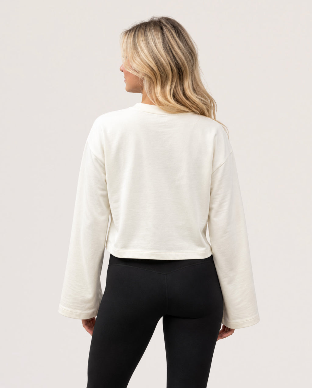 A woman shown from the back wearing a cream-colored long-sleeve cropped sweatshirt and black leggings against a plain light background.