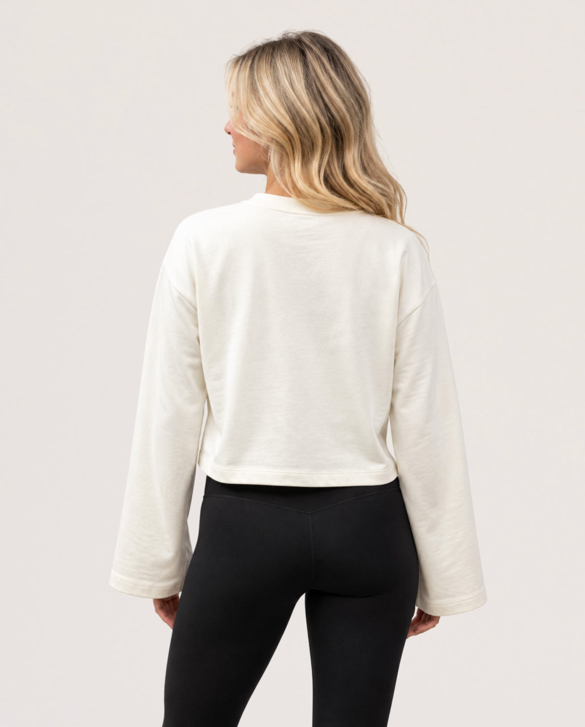 A woman shown from the back wearing a cream-colored long-sleeve cropped sweatshirt and black leggings against a plain light background.