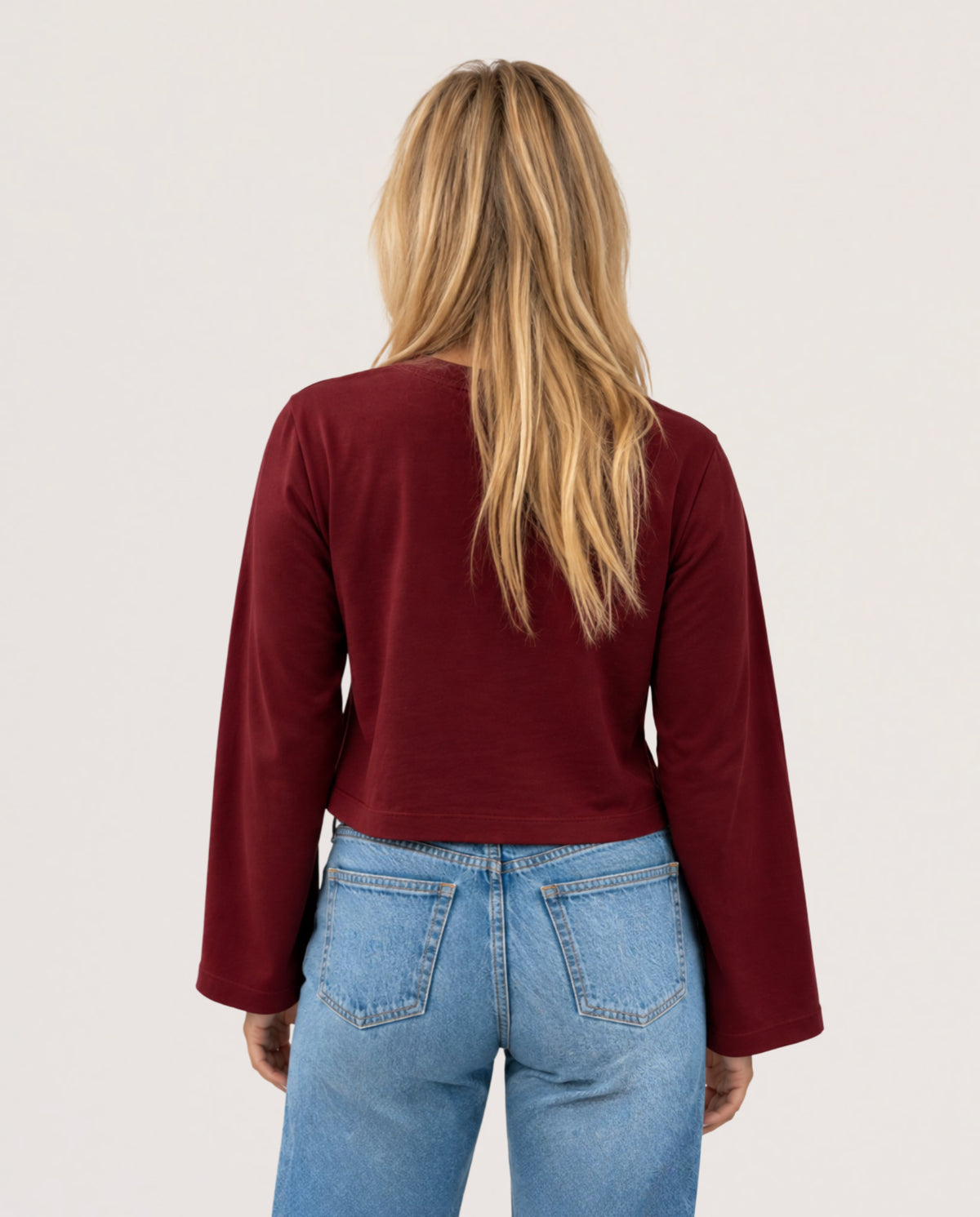 A woman viewed from behind wearing a long-sleeve cropped top in deep burgundy and light blue jeans, against a plain off-white studio background.