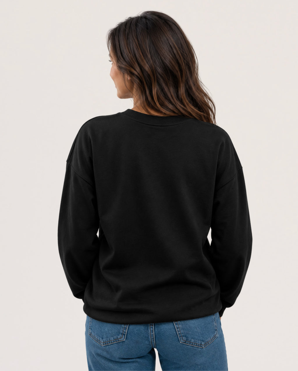 A woman viewed from the back wearing a plain black crewneck sweatshirt and blue jeans against a light off-white studio background.