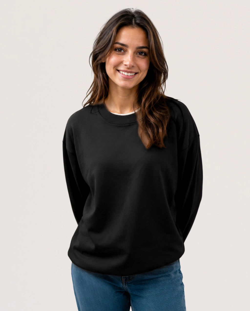 A woman with long brown hair smiling, wearing a plain black long-sleeve crewneck sweatshirt and blue jeans, photographed against a solid light gray background.