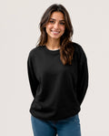 A woman with long brown hair smiling, wearing a plain black long-sleeve crewneck sweatshirt and blue jeans, photographed against a solid light gray background.