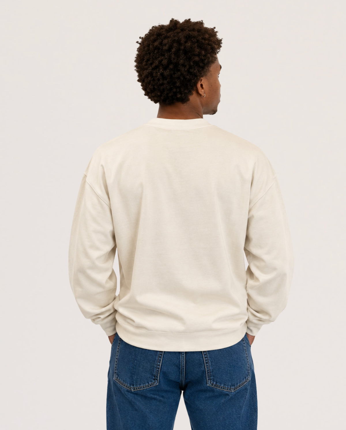 A man viewed from behind wearing a cream-colored crewneck sweatshirt and blue jeans against a plain light gray studio background.