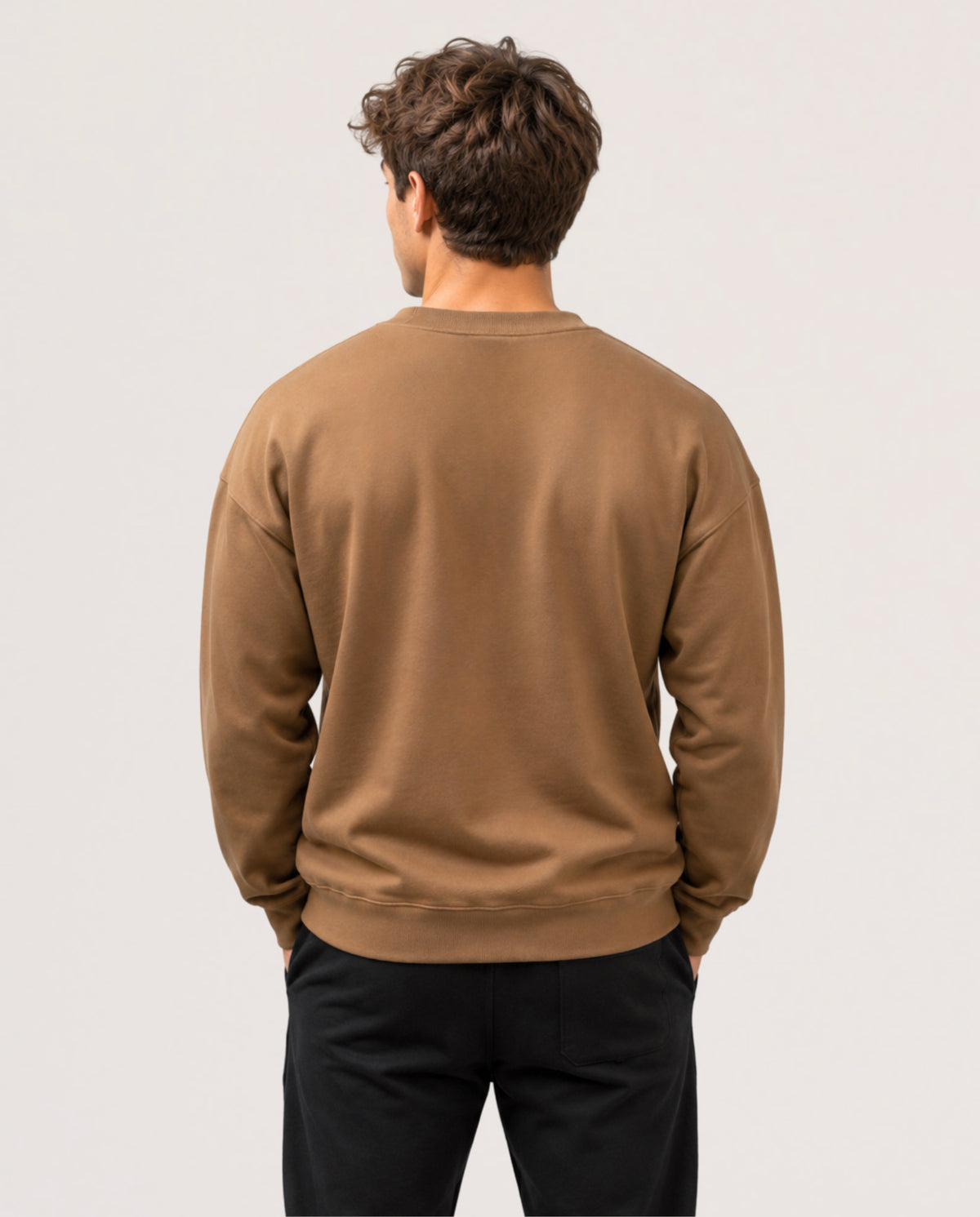 A man viewed from behind wearing a plain brown crewneck sweatshirt and black pants, standing against a light gray studio background.