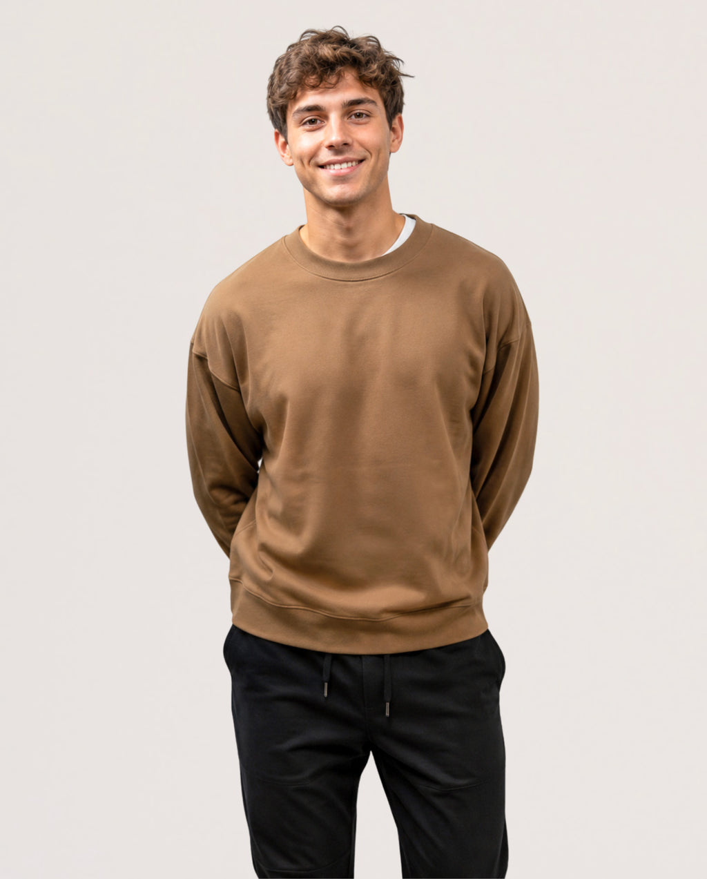 A smiling young man with curly brown hair wearing a brown crewneck sweatshirt and black sweatpants, standing against a light gray studio background.