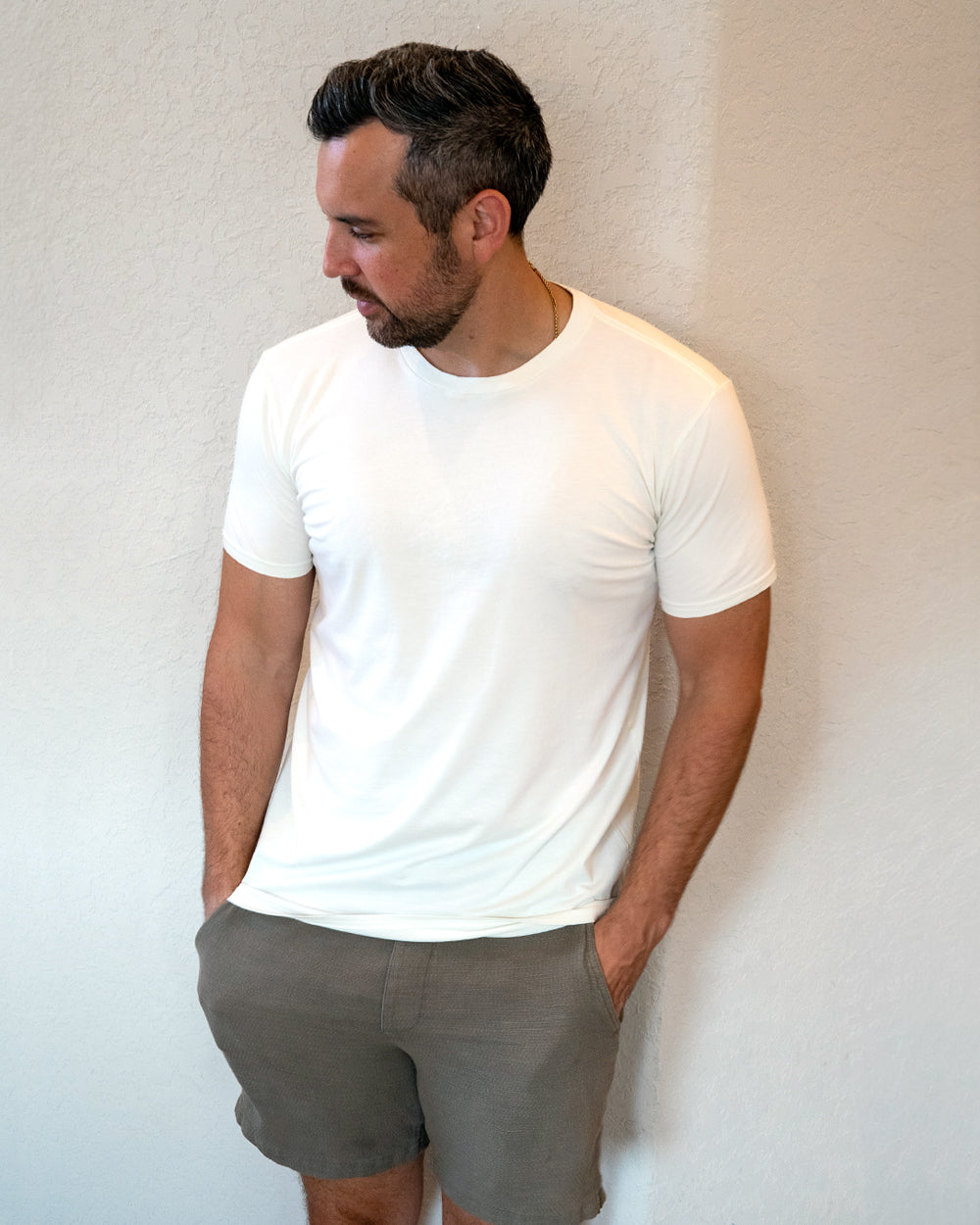 A man wearing a plain white short-sleeve crewneck t-shirt and gray shorts, standing against a textured off-white wall.