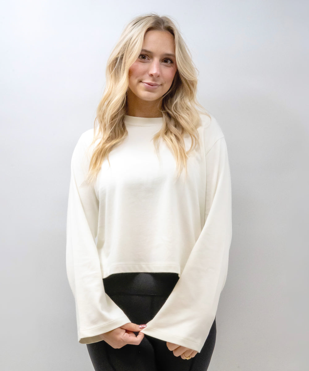 A blonde woman wearing a cream-colored long-sleeve crop top with flared bell sleeves and black pants, standing against a light gray studio background.