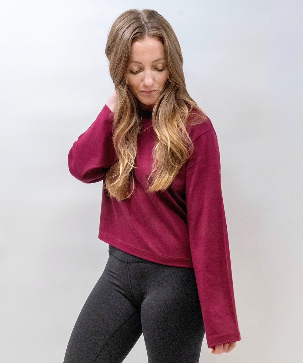 A woman with long blonde hair wearing a long-sleeve crewneck sweatshirt in deep burgundy and black leggings, posing against a light gray studio background.